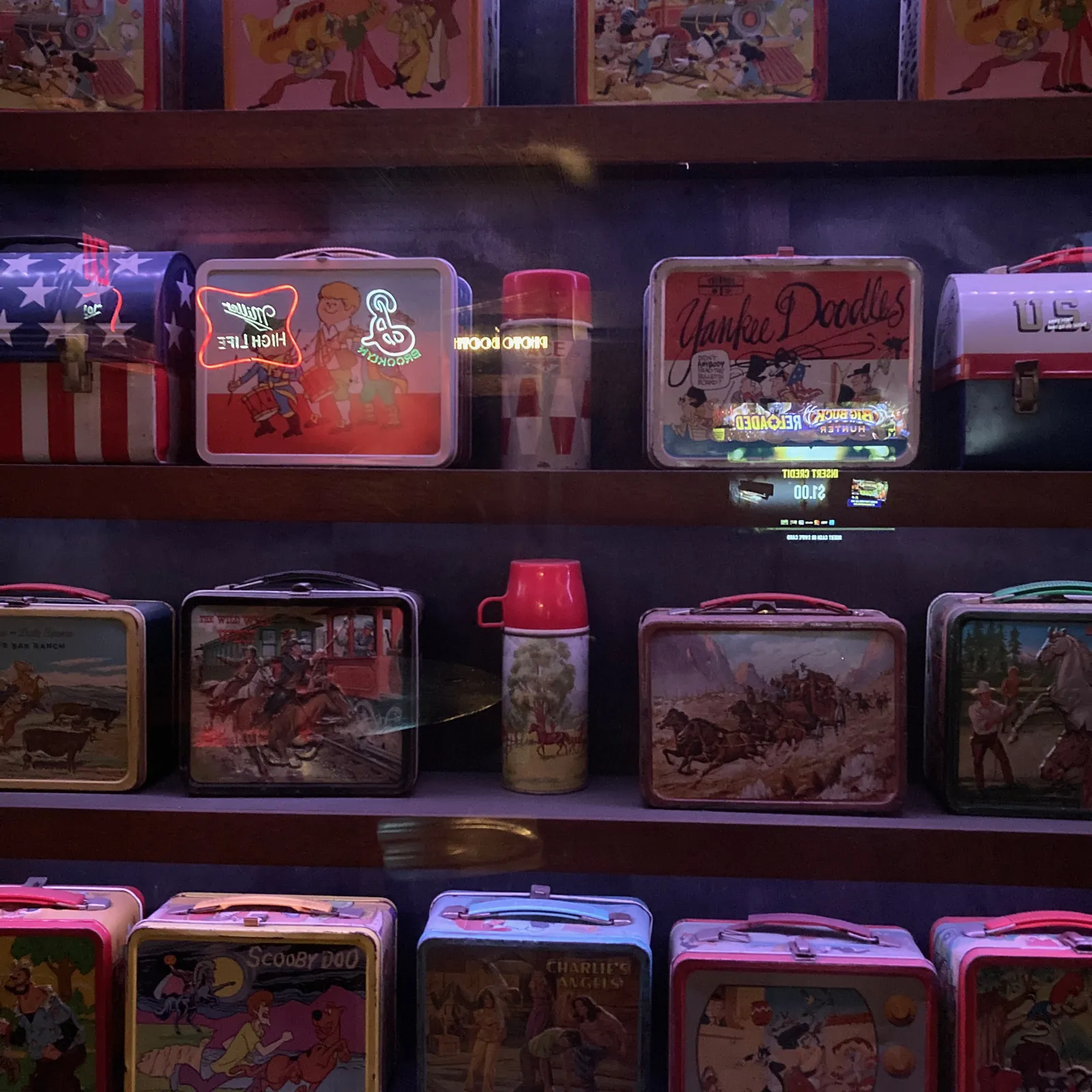 A wall of vintage metal lunchboxes and thermoses on shelves at Ace Bar, lit by neon beer signs — Yankee Doodles, Scooby-Doo, Charlie's Angels, and Western scenes among them.